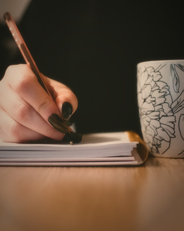 Soft hands writing in a notebook beside a floral mug, in warm low light